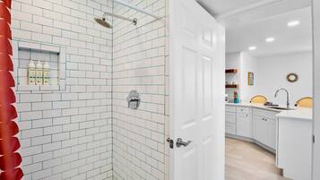 Un baño moderno con azulejos blancos tipo metro, una ducha con cabezal de ducha manual y una puerta blanca que da a una zona de cocina.