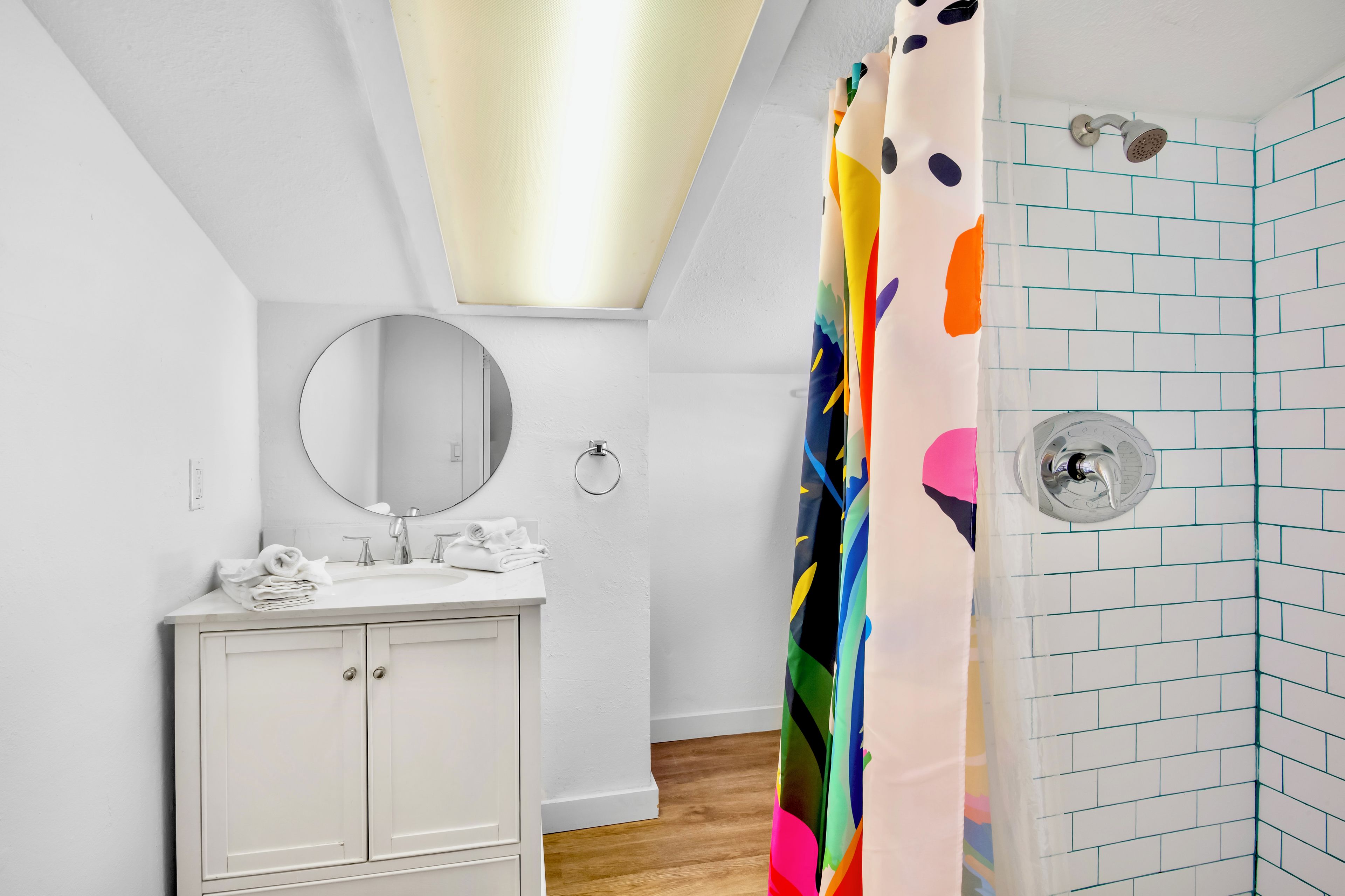 Un baño con tocador blanco, un espejo redondo, ducha con cortina colorida y pared con azulejos.