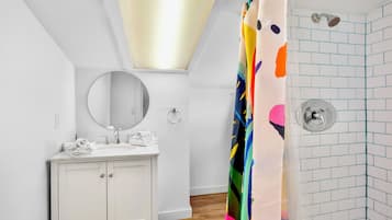 Un baño con tocador blanco, un espejo redondo, ducha con cortina colorida y pared con azulejos.
