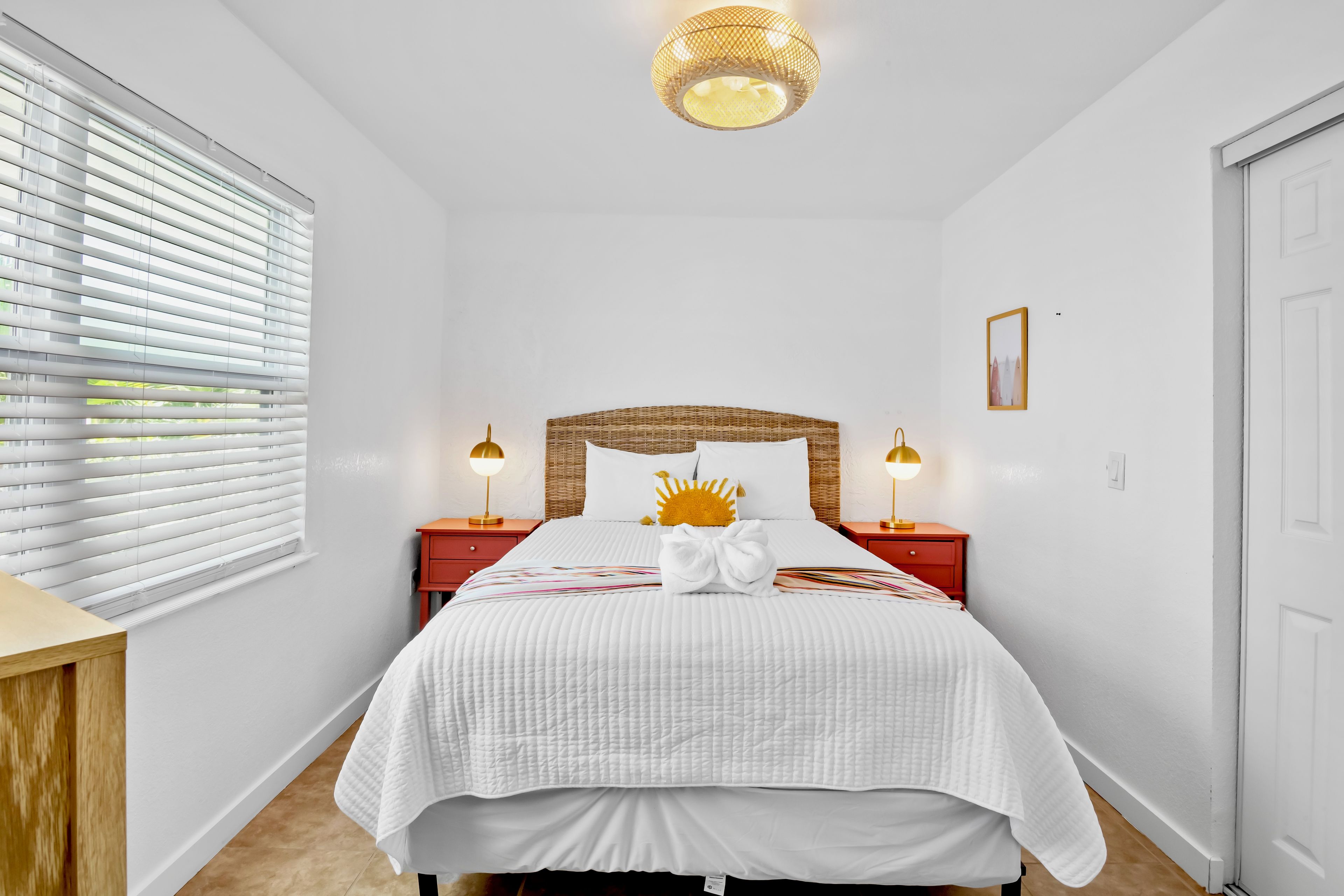 Un dormitorio con una cama, dos mesitas de noche, un aplique de pared y una ventana con persianas.