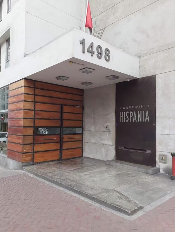 L’entrée d’un immeuble, avec le grand numéro « 1498 » au-dessus de la porte, une enseigne indiquant « CONDOMINIO HISPANIA » et un drapeau rouge flottant au-dessus de l’entrée.
