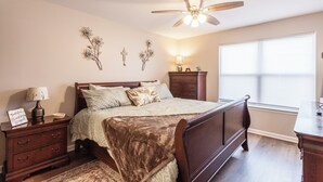 Quarto com cama, mesas de cabeceira, cômoda, ventilador de teto, obras de arte na parede e janela com persianas.