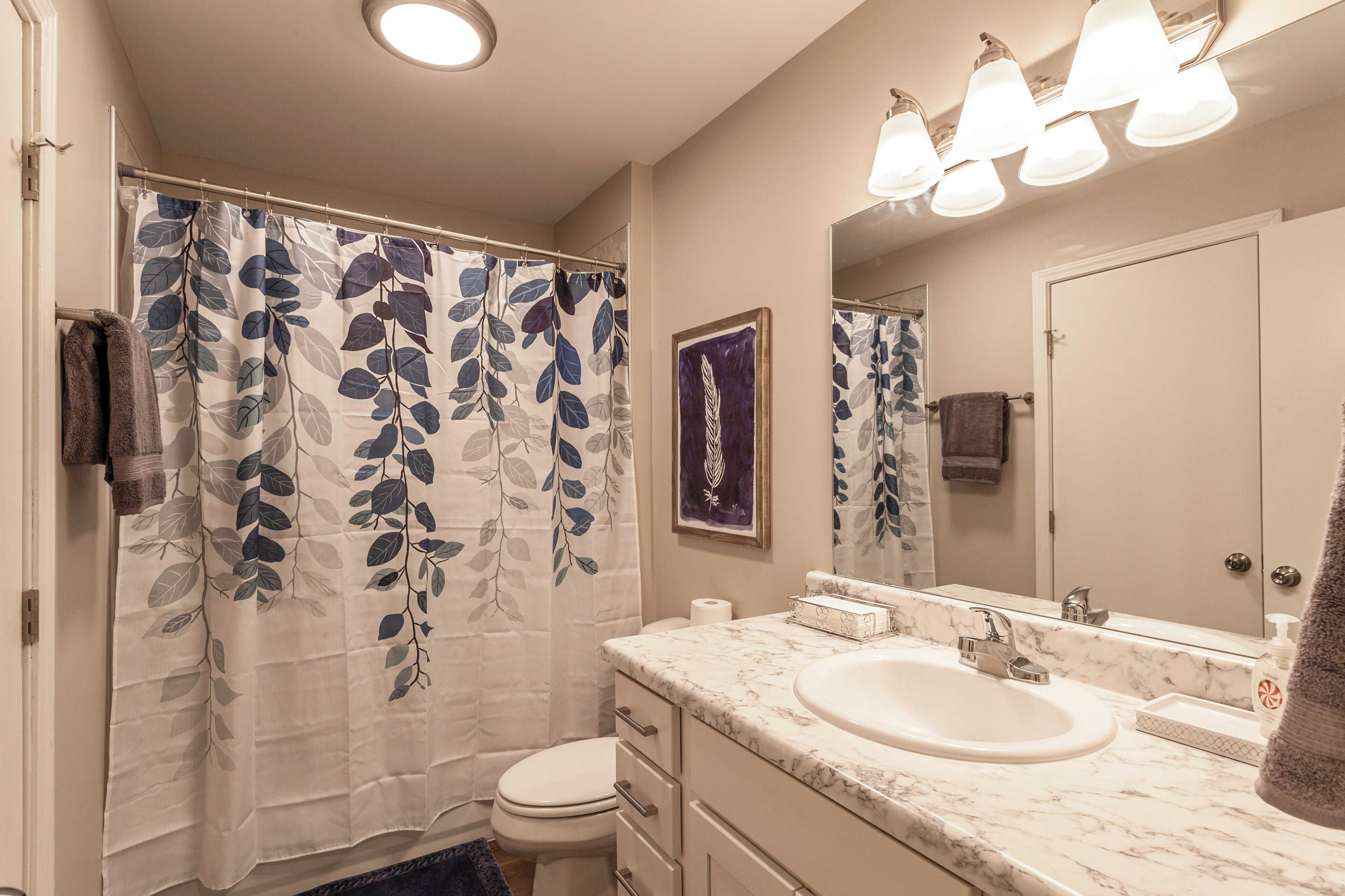 A bathroom with a shower curtain featuring a leaf pattern, a toilet, a sink with a marble countertop, and a mirror above the sink.