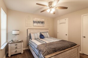 Ein Schlafzimmer mit hölzernem Kopfteil, einem Bett mit gemusterten Textilien, einem Nachttisch mit Lampe, einem Deckenventilator und Kunstwerken an den Wänden.