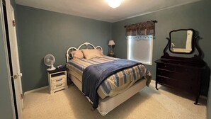 A bedroom with a bed, bedside table, lamp, mirror, dresser, and a window with curtains.