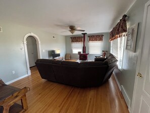 A living room with a dark-colored sofa, a wooden coffee table, a ceiling fan, a red wood-burning stove, and a television.