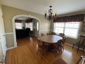 A dining room with a round wooden table surrounded by chairs, a chandelier, and a view of a living area through an archway.