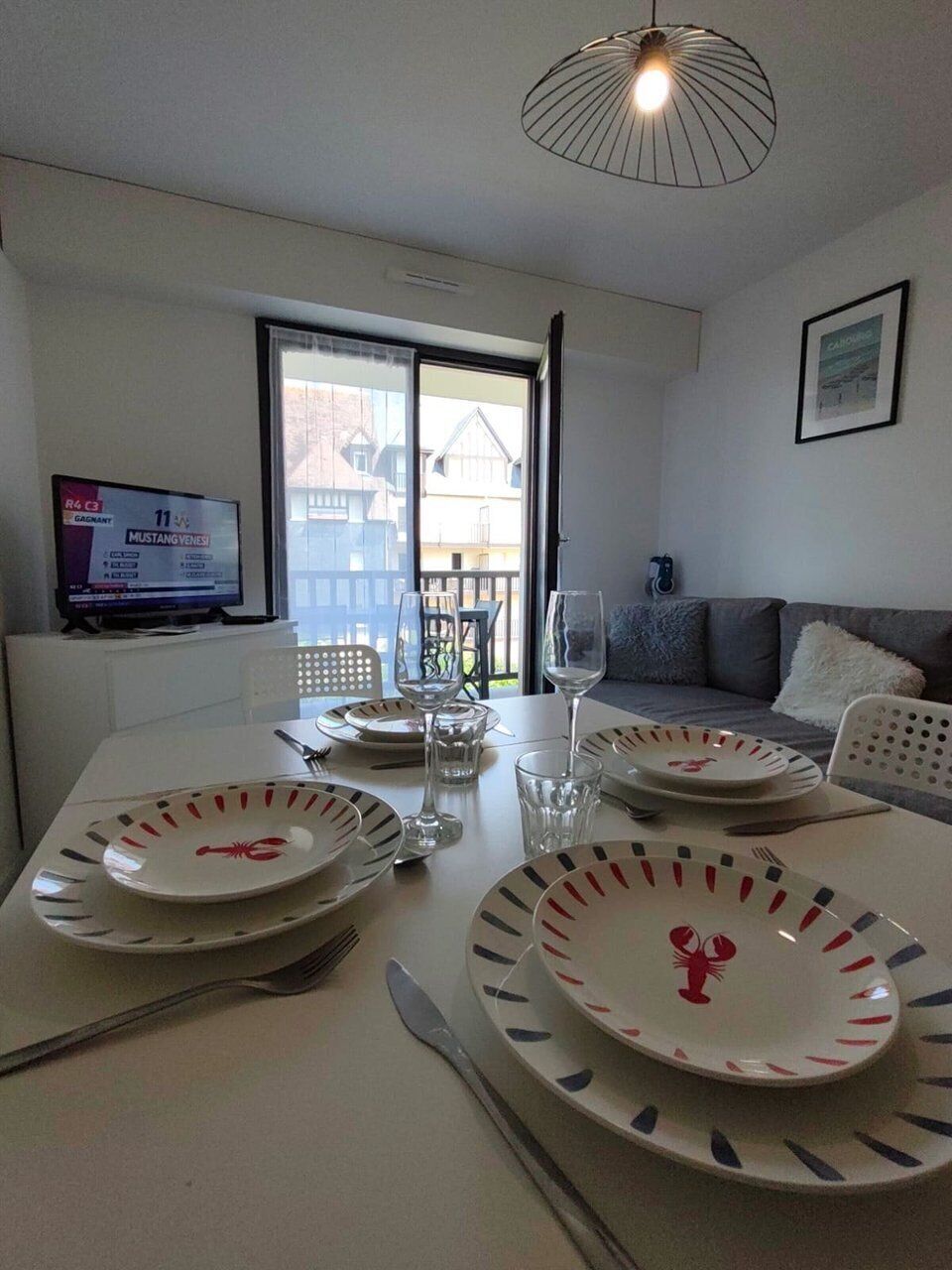 Een eethoek met een tafel gedekt voor twee personen, een televisie waarop een sportevenement wordt vertoond, en een balkon met buitenmeubels.