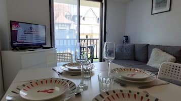 Een eethoek met een tafel gedekt voor twee personen, een televisie waarop een sportevenement wordt vertoond, en een balkon met buitenmeubels.
