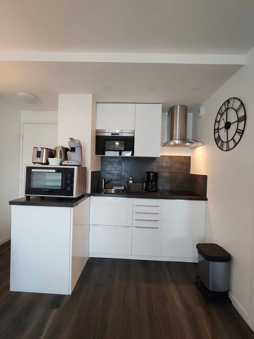 A compact kitchen with white cabinets, a microwave, a coffee maker, and a clock on the wall.