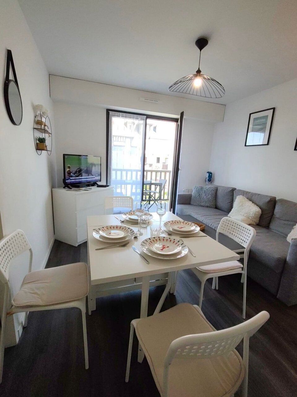 Un coin repas avec une table dressée pour quatre personnes, des chaises blanches, un canapé gris et un téléviseur posé sur un meuble blanc.