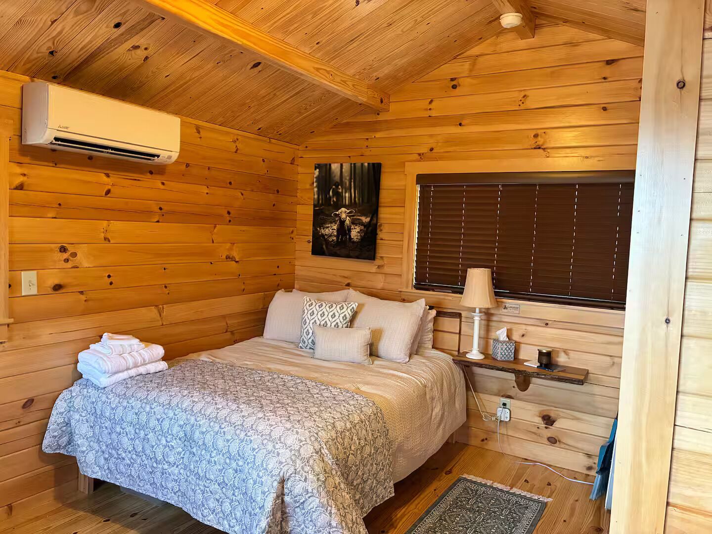 Un dormitorio de cabaña de madera con una cama, almohadas, una lámpara de noche y una ventana con persianas.