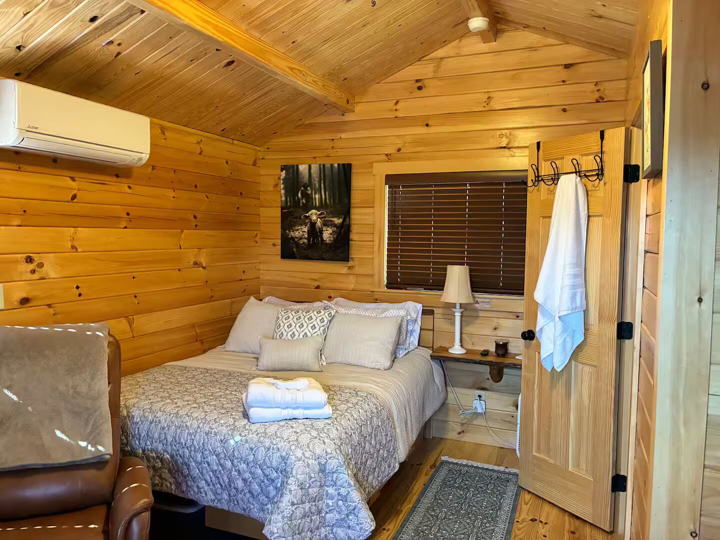 Un dormitorio rústico de cabaña de madera con una cama, una silla, una mesita de noche y una ventana con persianas.