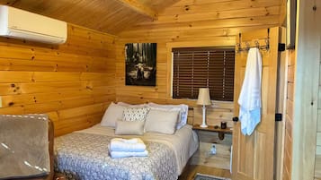Un dormitorio rústico de cabaña de madera con una cama, una silla, una mesita de noche y una ventana con persianas.