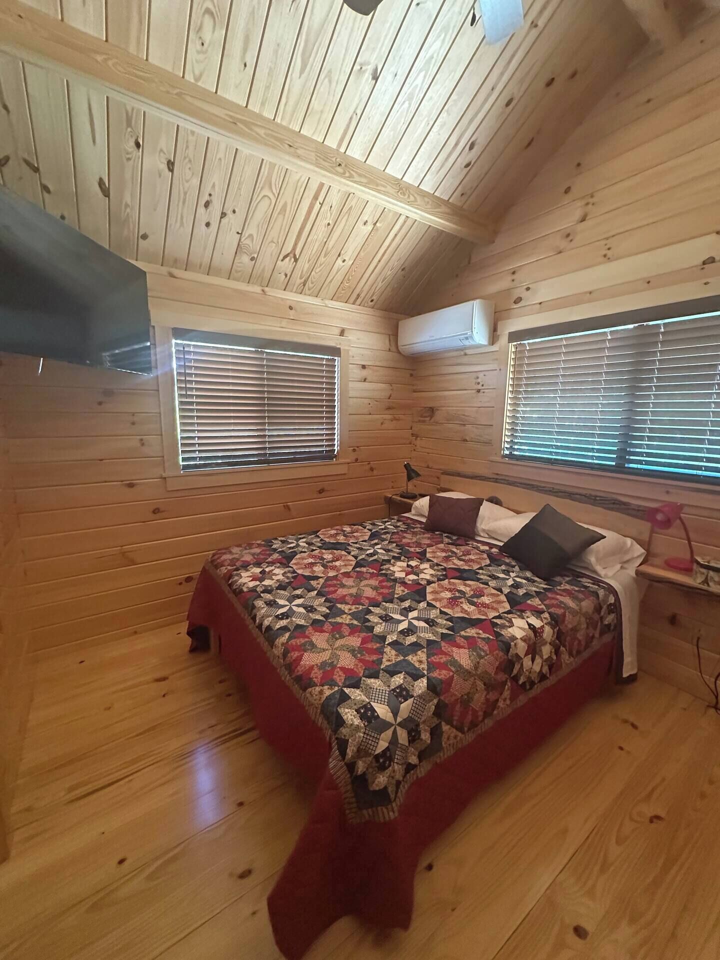 A bedroom with a wooden ceiling, two windows with blinds, a bed with a quilt, and an air conditioning unit.
