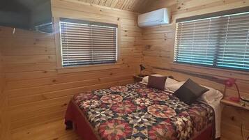 A bedroom with a wooden ceiling, two windows with blinds, a bed with a quilt, and an air conditioning unit.