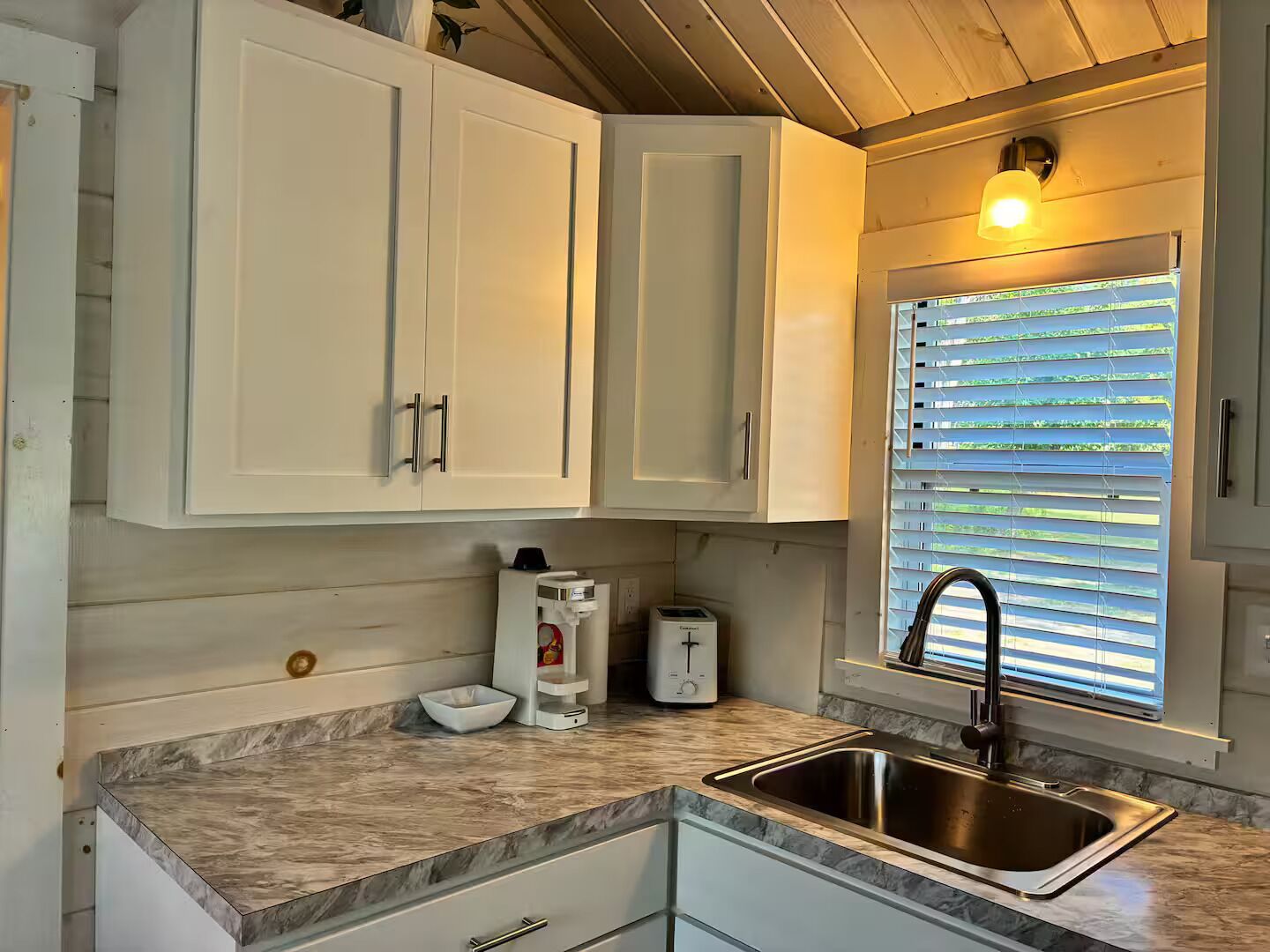 A kitchen with white cabinets, a stainless steel sink, a window with blinds, and a countertop with a coffee maker and toaster.