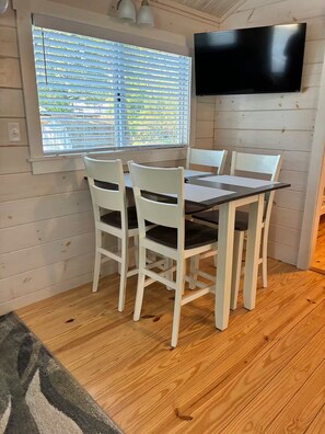 Een eetruimte met een tafel en stoelen, een raam met zonwering en een flatscreen televisie aan de muur.