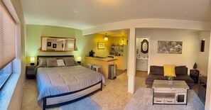 A bedroom with a bed, bedside tables, a desk, and a chair. There is a kitchenette with a microwave, toaster, and sink. The room has a large window and a door leading to a living area with a sofa and a coffee table. - Trail Head Studio Suite (Village of Oak Creek)