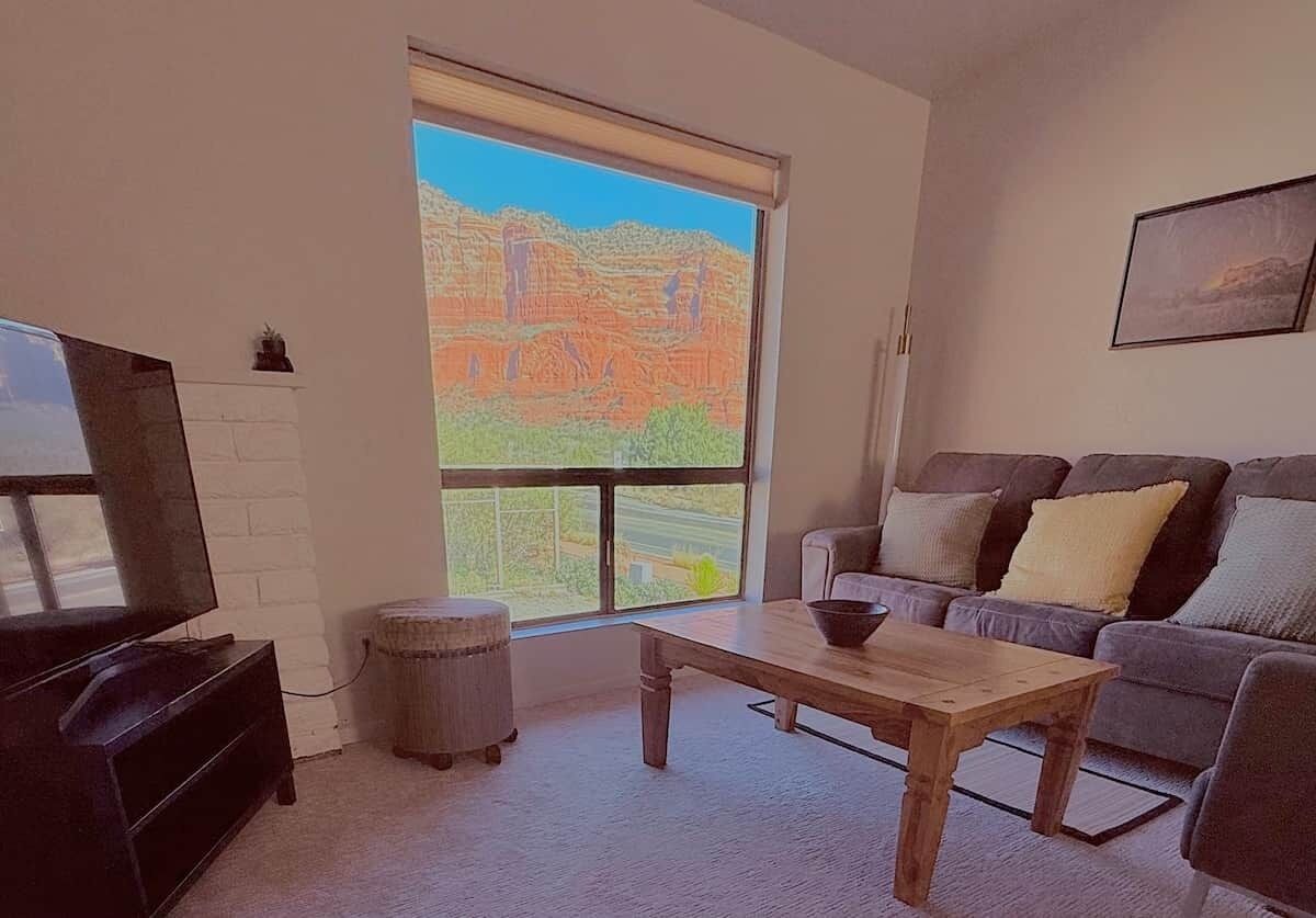 A living room with a television, a sofa, a coffee table, and a large window with a view of a landscape.