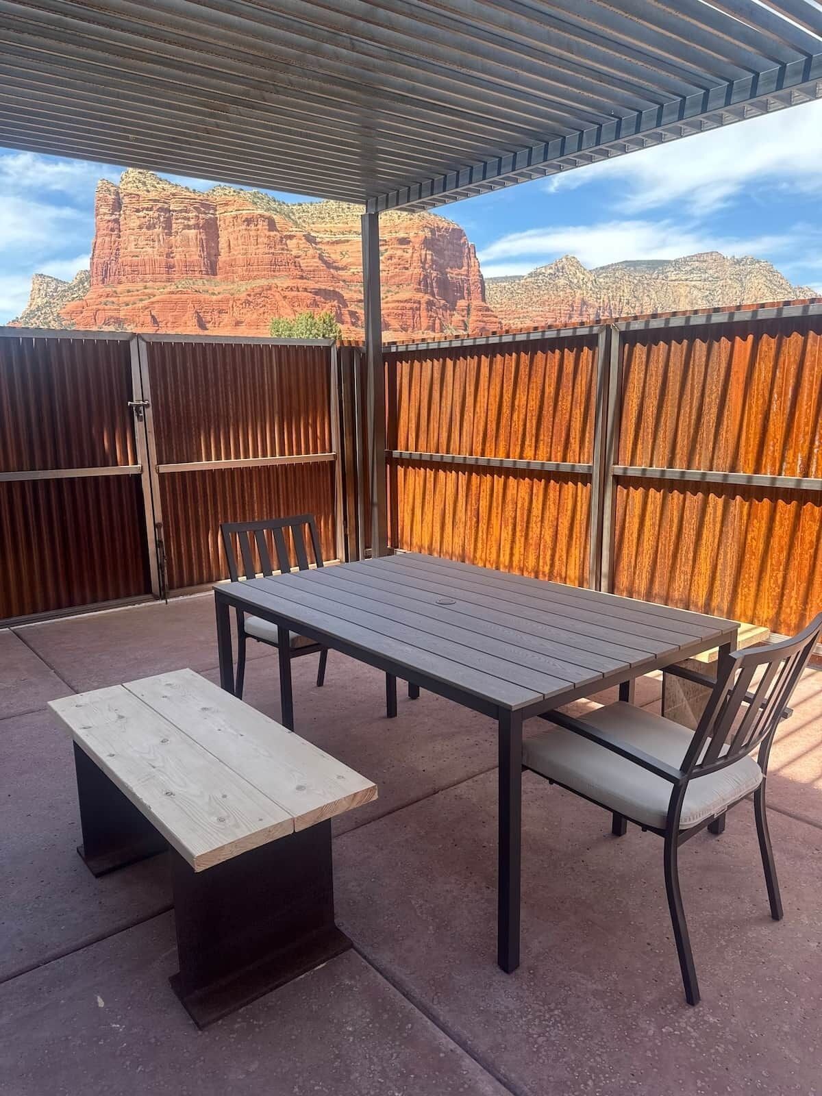 Un patio al aire libre con una mesa y sillas, un banco de madera y una valla metálica.