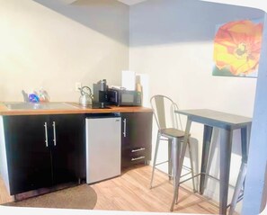 Eine kompakte Küchenzeile mit Kühlschrank, Mikrowelle und Spüle. Ein kleiner Esstisch mit zwei Stühlen steht bereit. Ein Gemälde ist an der Wand sichtbar.