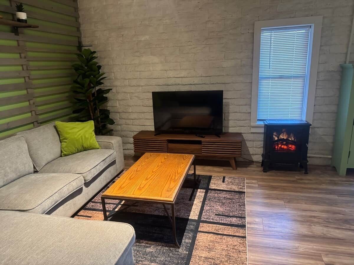Ein Wohnzimmer mit einer Couch, einem Holztisch, einem Flachbildfernseher, einem Kamin und einem Fenster mit Jalousien.