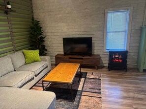 Ein Wohnzimmer mit einer Couch, einem Holztisch, einem Flachbildfernseher, einem Kamin und einem Fenster mit Jalousien.