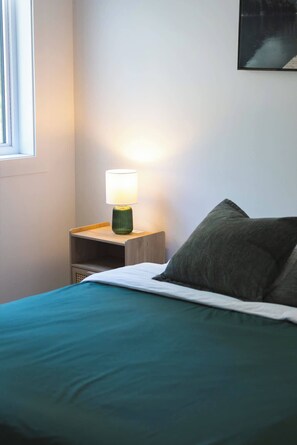 A bed with a teal bedspread and a pillow, a bedside table with a lamp, and a window with blinds. - Nature Getaway & Spa | Conifer (Notre-Dame-de-la-Merci)