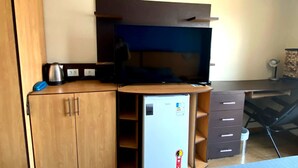 Une chambre de taille modeste, équipée d’une télévision, d’un petit réfrigérateur, d’un bureau et d’une chaise.