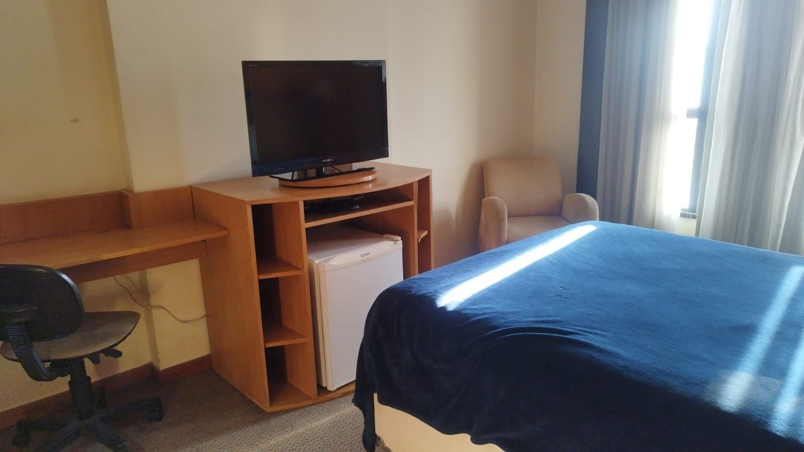 Una habitación de hotel con una cama, un escritorio con computadora, un televisor y un refrigerador pequeño.