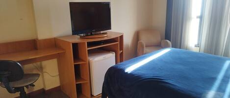Una habitación de hotel con una cama, un escritorio con computadora, un televisor y un refrigerador pequeño.