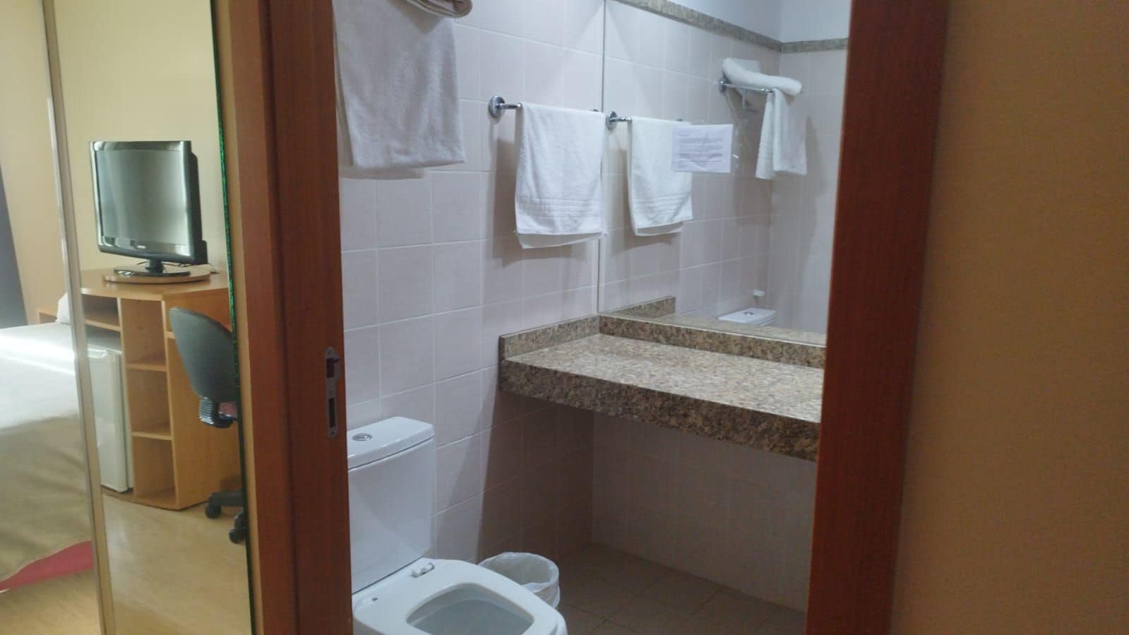 Een hotelbadkamer met toilet, wastafel en een spiegel. In de aangrenzende kamer staat een houten bureau en een stoel.