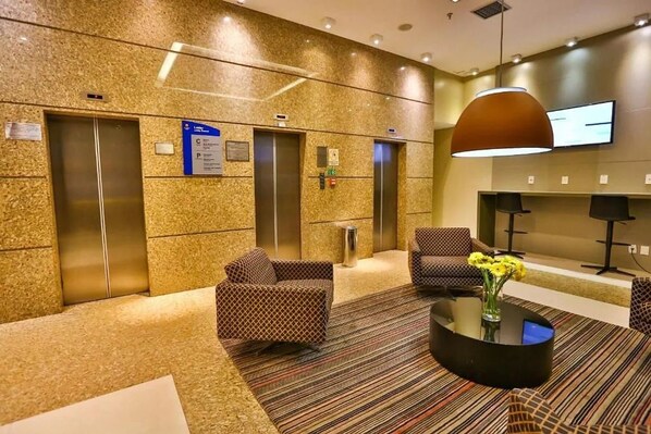 Un hall d'entrée doté de deux ascenseurs, de canapés à motifs, d'une table basse avec un vase et d'un téléviseur fixé au mur.