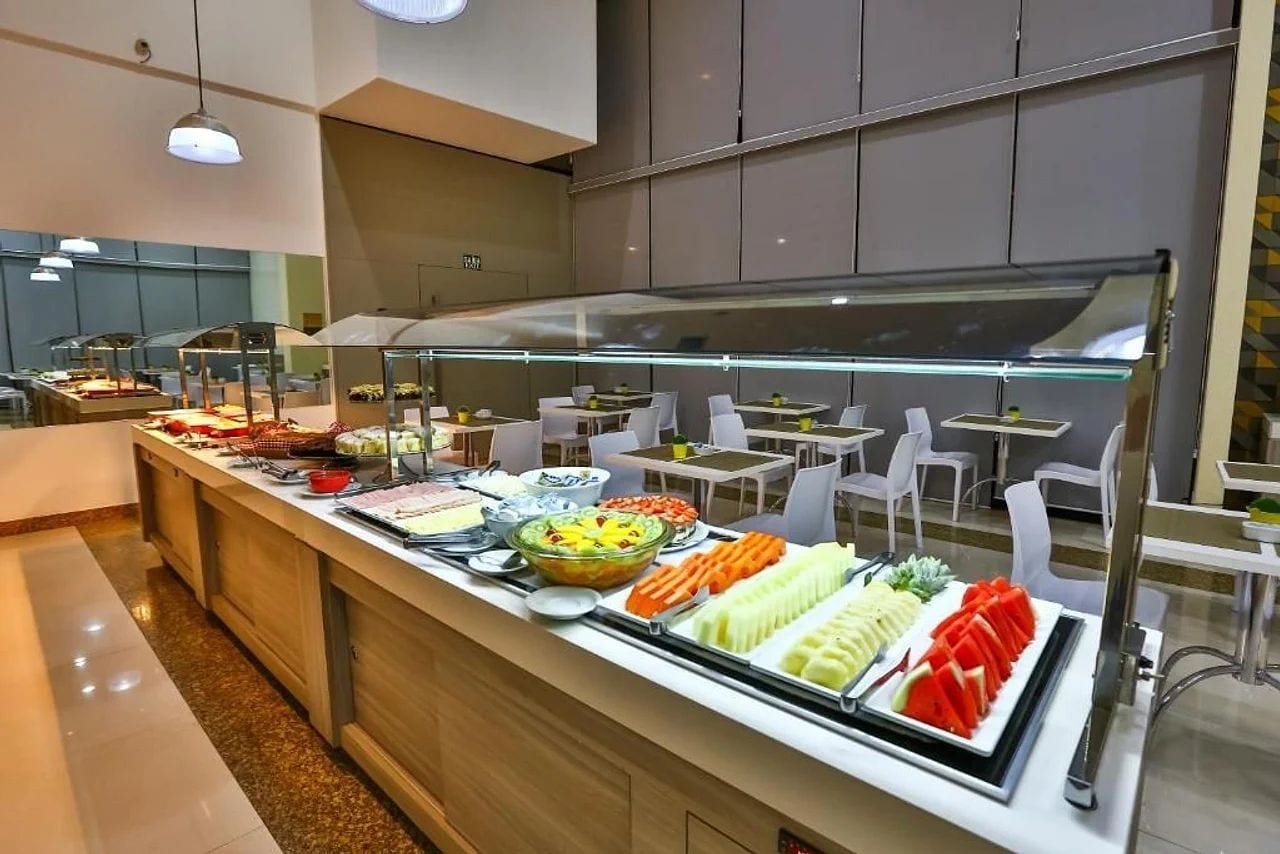 A buffet area with various food options, including fruits, vegetables, and prepared dishes, arranged on a counter with seating in the background.