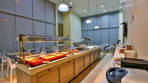 Un espace buffet avec plusieurs postes de service pour les différents plats, un espace repas avec des tables et des chaises, et un comptoir avec une caisse enregistreuse.