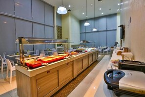 Un espace buffet avec plusieurs postes de service pour les différents plats, un espace repas avec des tables et des chaises, et un comptoir avec une caisse enregistreuse.