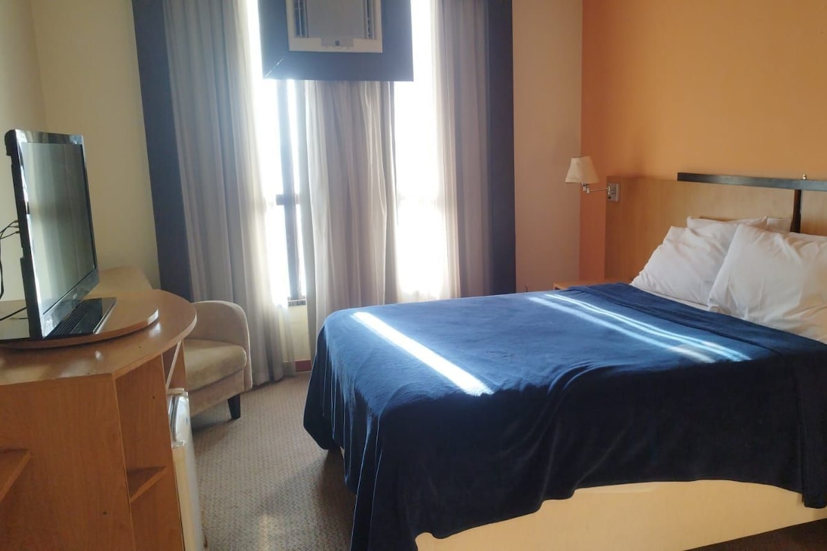 Une chambre d’hôtel comprenant un lit, un bureau avec un ordinateur portable, une chaise, une table de chevet et une fenêtre avec des rideaux.