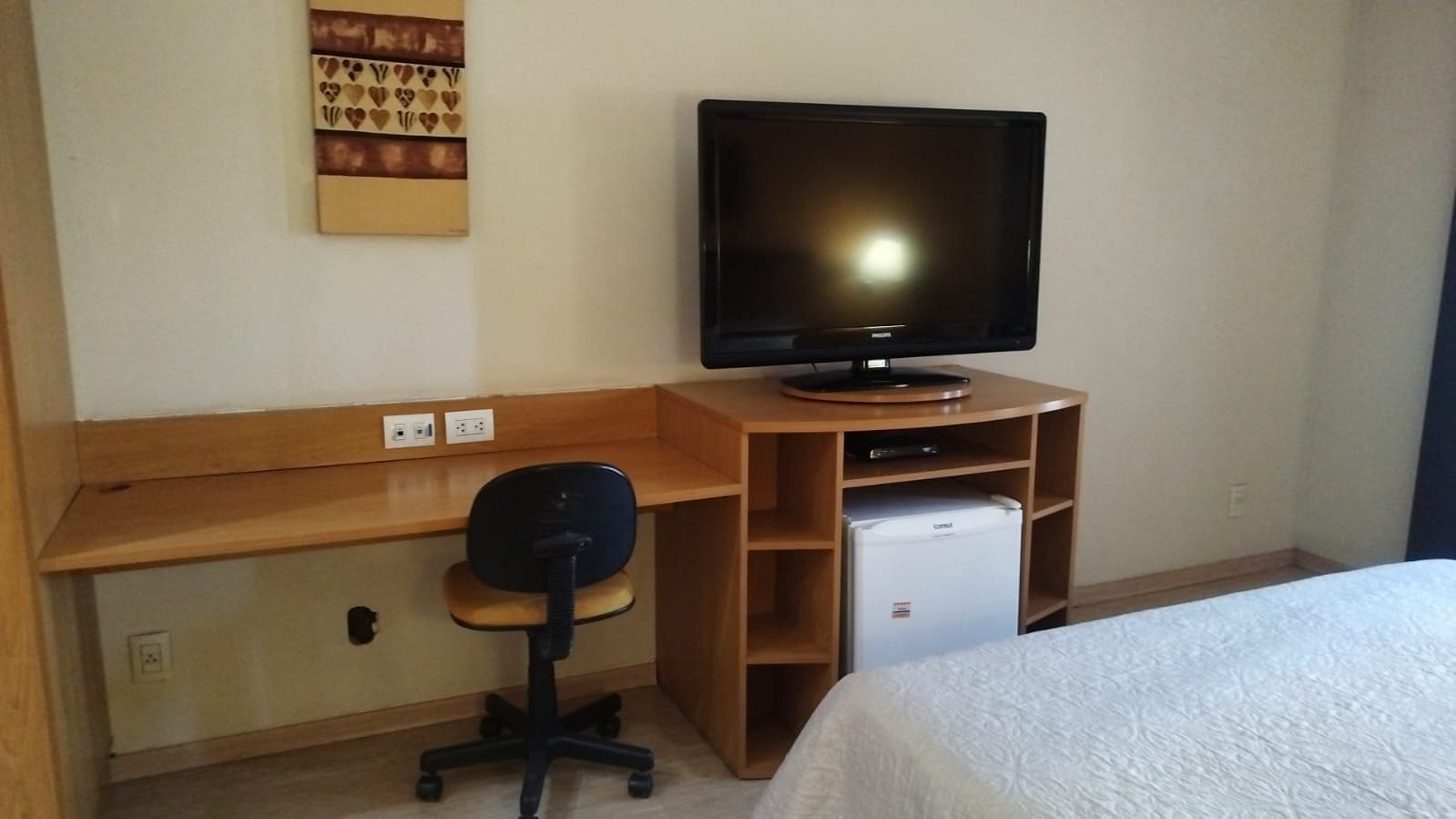 Ett hotellrum med en säng, ett skrivbord med en dator, en stol, en tv och ett minikylskåp.