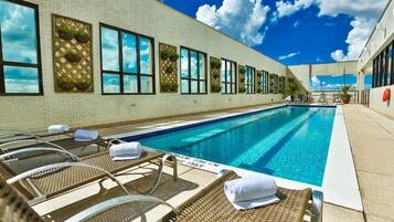 Una piscina con sillones de descanso y tumbonas, cielo despejado y un edificio de ladrillo con ventanas.