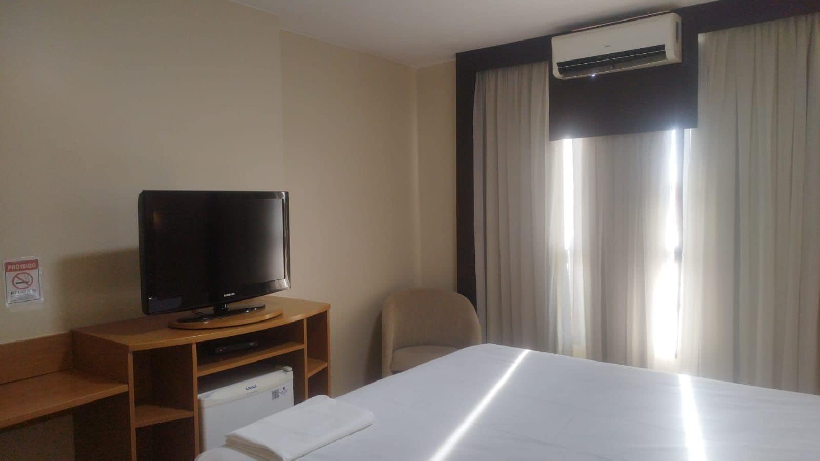 Una camera d'albergo con un letto, una televisione su un supporto in legno, una sedia e un condizionatore d'aria.