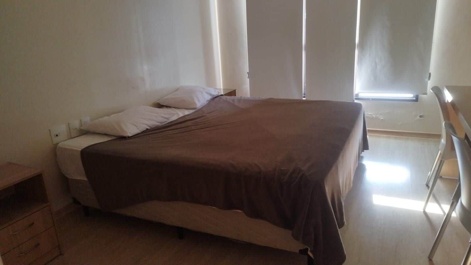 A single bed with a brown bedspread, a nightstand, and a window with blinds.
