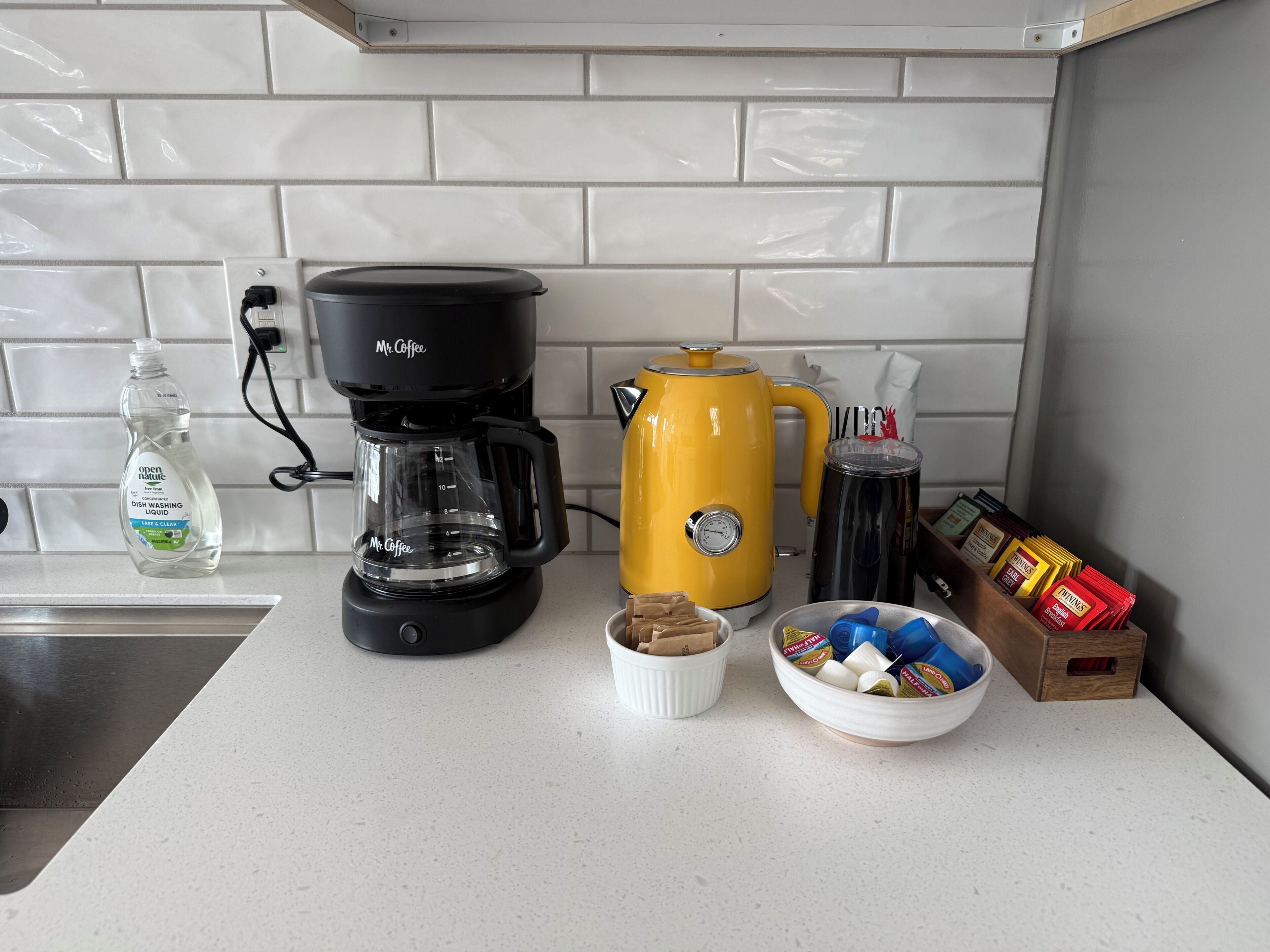 Una encimera de cocina con una cafetera, un hervidor amarillo, una taza, una botella de jabón para lavar trastes y un tazón de bocadillos.