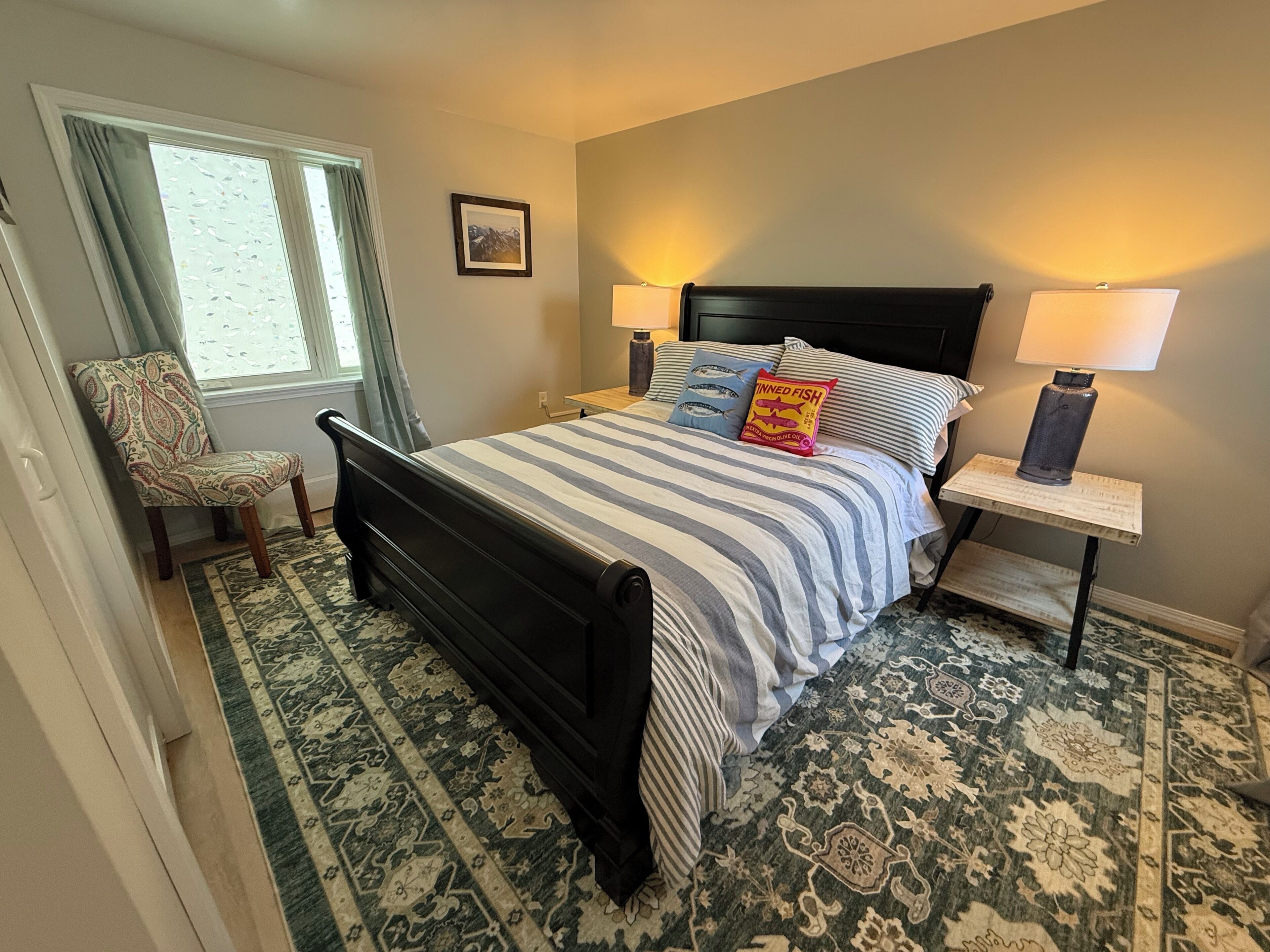 A bedroom with a bed, two bedside tables, a chair, a window with curtains, and a patterned rug.