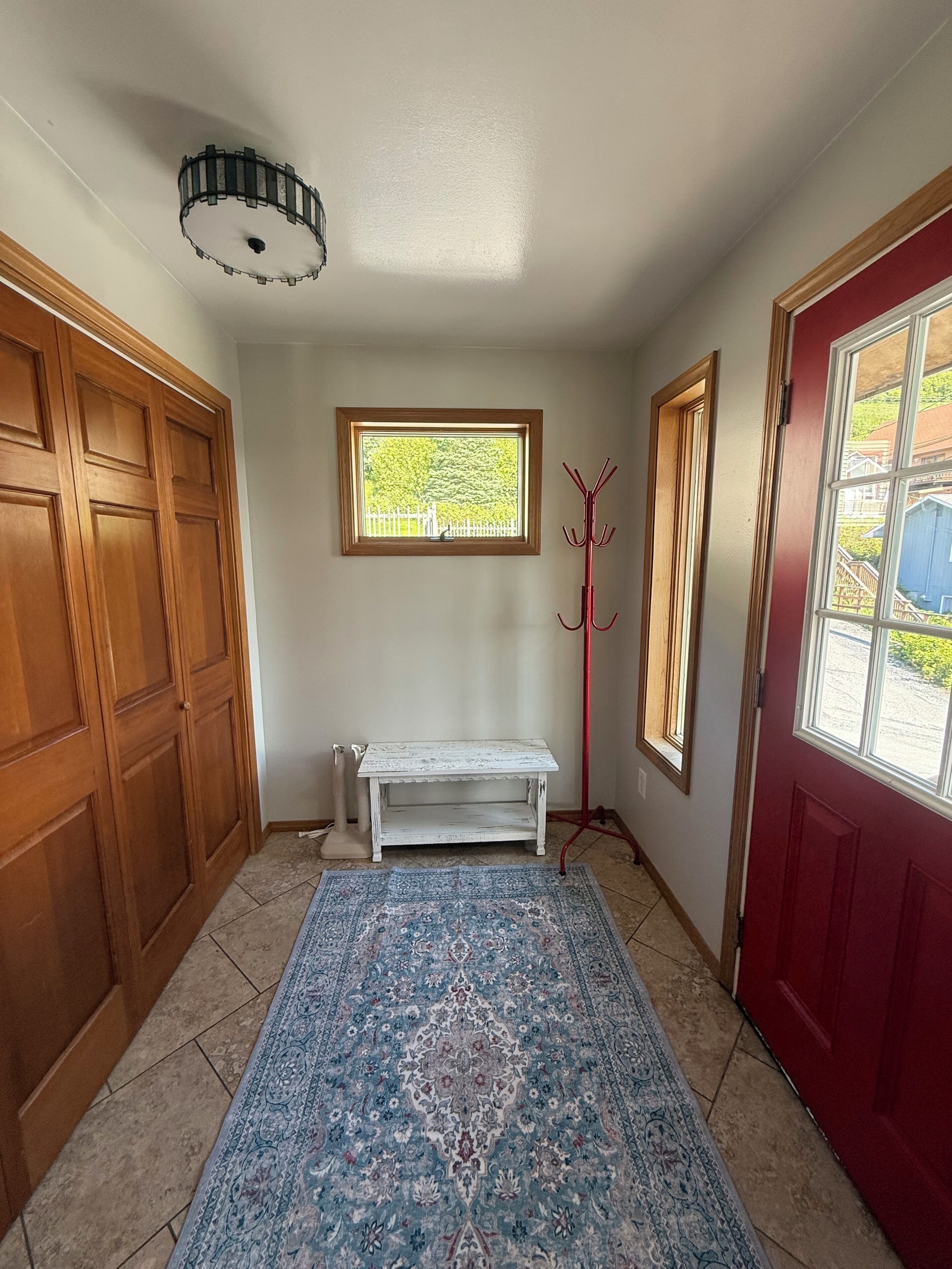 A hallway with a wooden door, a red door, a bench, a coat rack, and a window with a view of greenery.