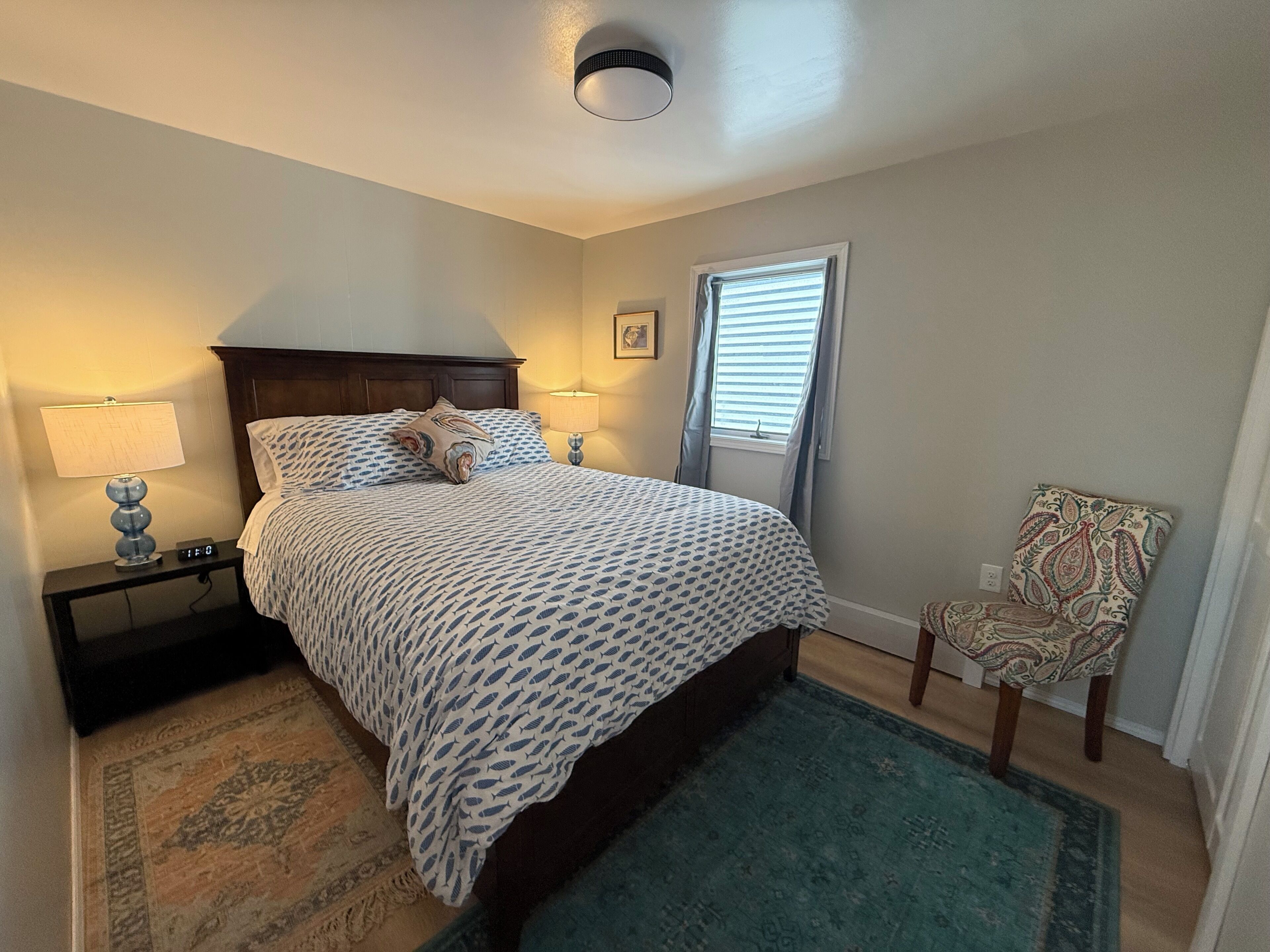 A bedroom with a bed, two bedside lamps, a chair, a window with blinds, and a rug.