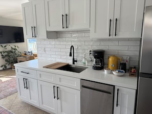 A modern kitchen with white cabinets, a stainless steel dishwasher, a black coffee maker, and a yellow kettle.