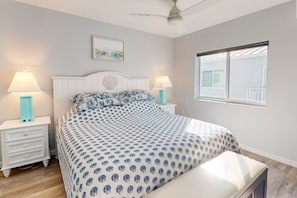Quarto com cama, duas mesas de cabeceira, um criado-mudo, ventoinha no teto, janela e um quadro na parede.
