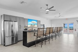 Cozinha moderna com geladeira de aço inoxidável, ilha central com cadeiras e vista para a praia pela porta.