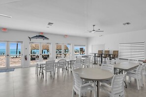 Um espaço amplo e interno com várias mesas e cadeiras, janelas grandes com vista para a praia, e um sinal de saída.